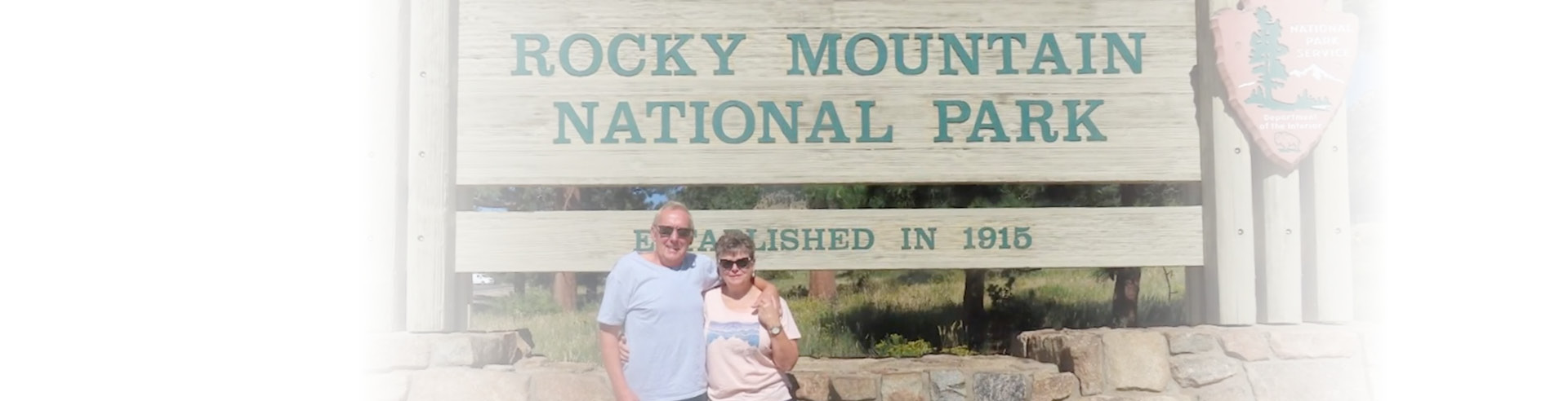 Two people including Billy Stryke standing, arms around each other in front of the a sign that reads "rock mountain national park"